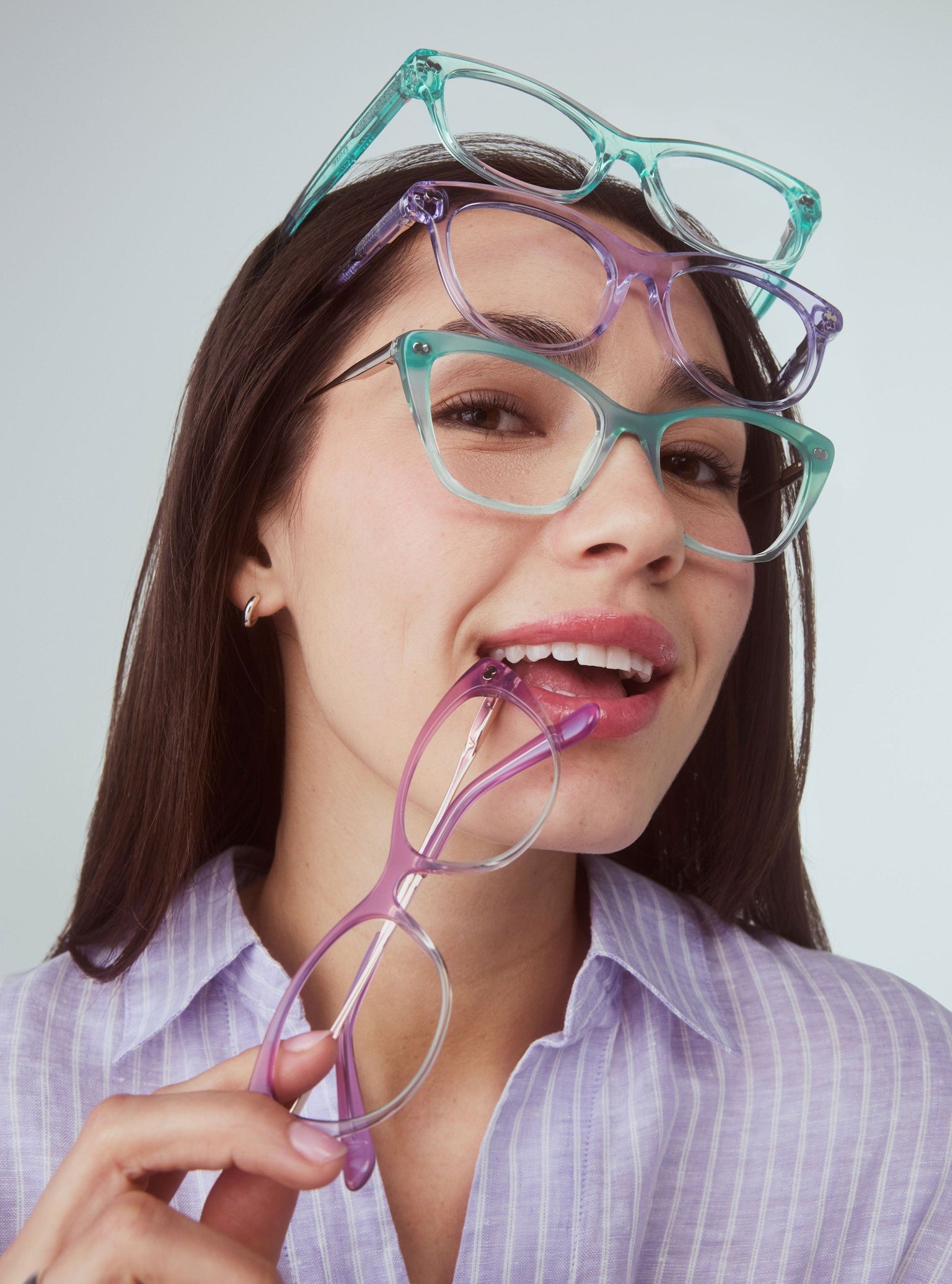 A woman wearing teal Pair glasses with three more colourful frames stacked on her head, holding a fourth to her mouth